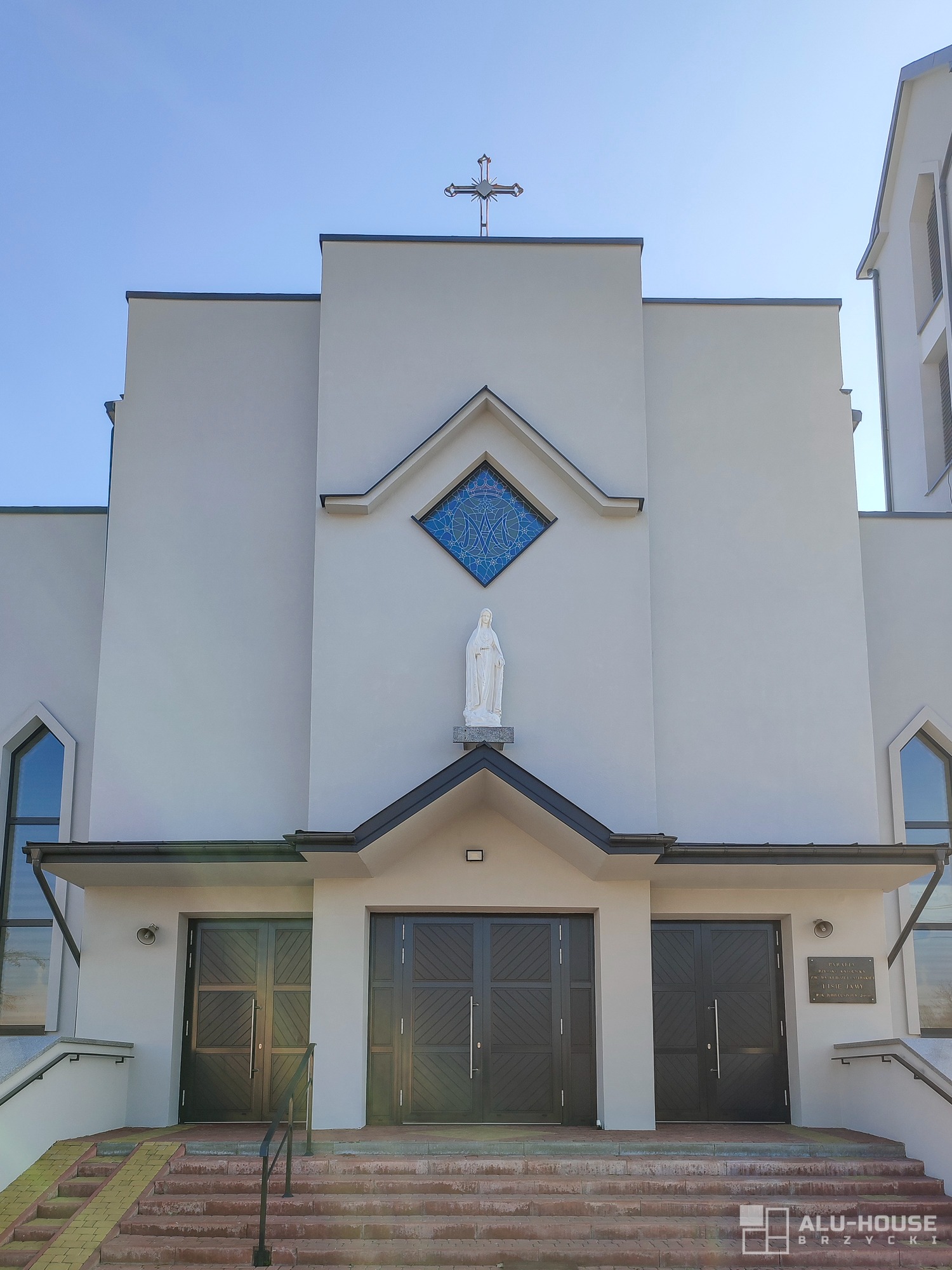 Fasada kościoła z trzema parami ciemnych drzwi zewnętrznych, nad środkowymi drzwiami statua Matki Boskiej i witraż w kształcie rombu, na szczycie krzyż.