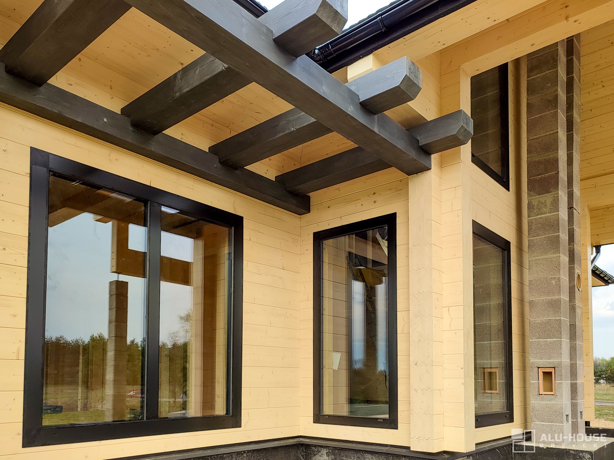 Nowoczesny dom z jasnego drewna z czarnymi ramami okiennymi, widoczne także ciemne drewniane belki stropowe i fragment murowanego komina.