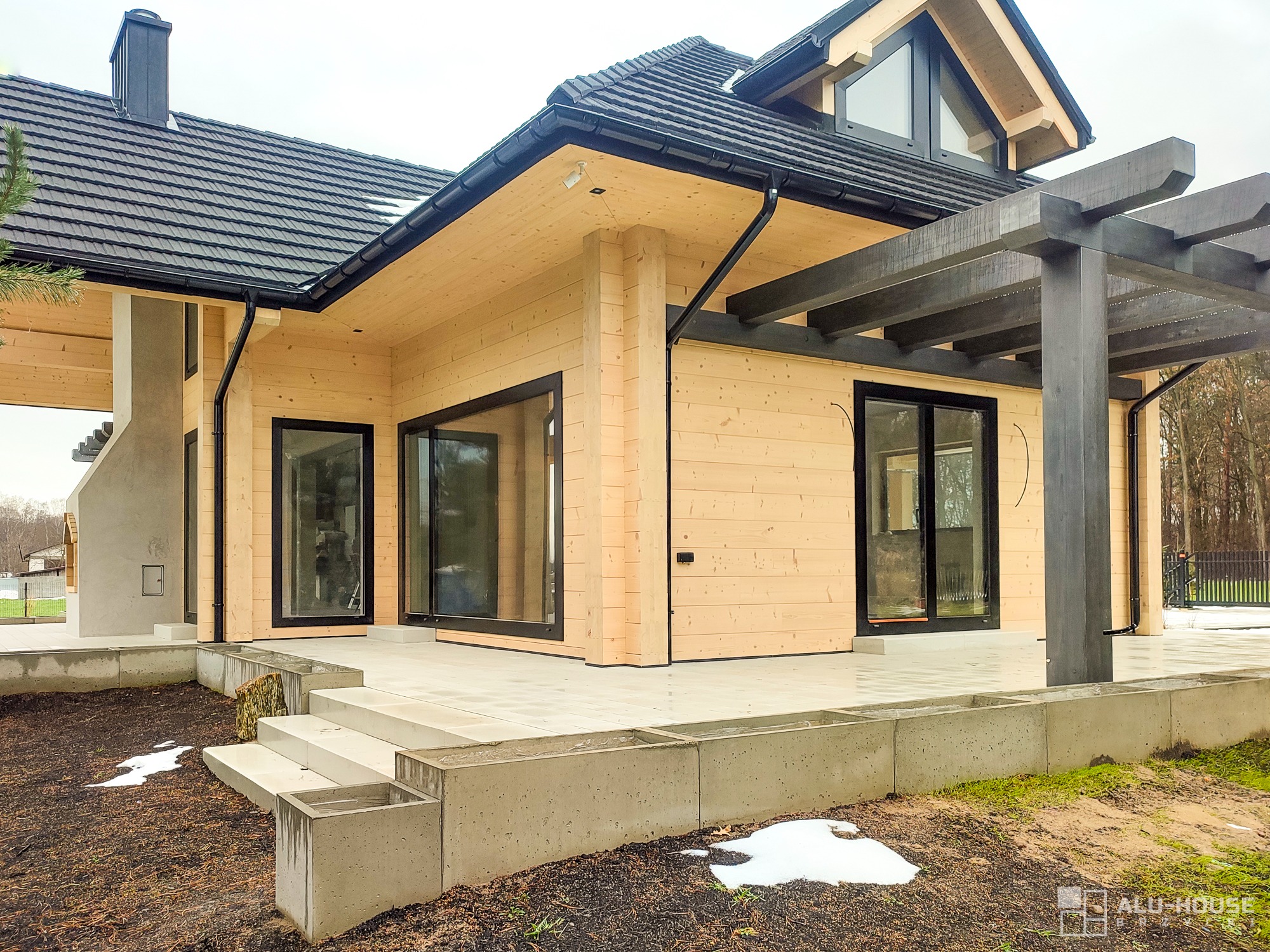 Nowoczesny dom z jasnego drewna z czarną dachówką, czarnymi ramami okien i przesuwnymi drzwiami, oraz ciemną drewnianą pergolą