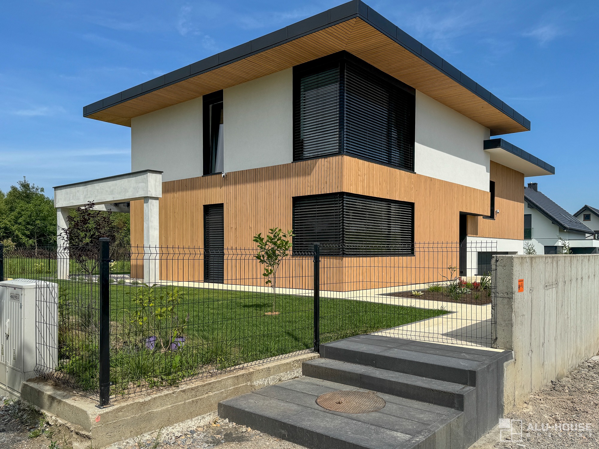 Nowoczesny dom z drewnianą elewacją, czarnymi ramami okiennymi i metalową siatką ogrodzeniową, widok z przodu, słoneczny dzień.