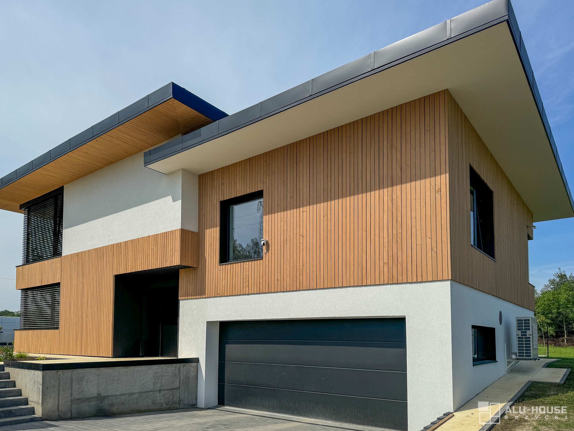 Nowoczesny dom z drewnianą elewacją, płaskim dachem i garażem z szarą bramą segmentową, widoczne okna z czarnymi ramami.