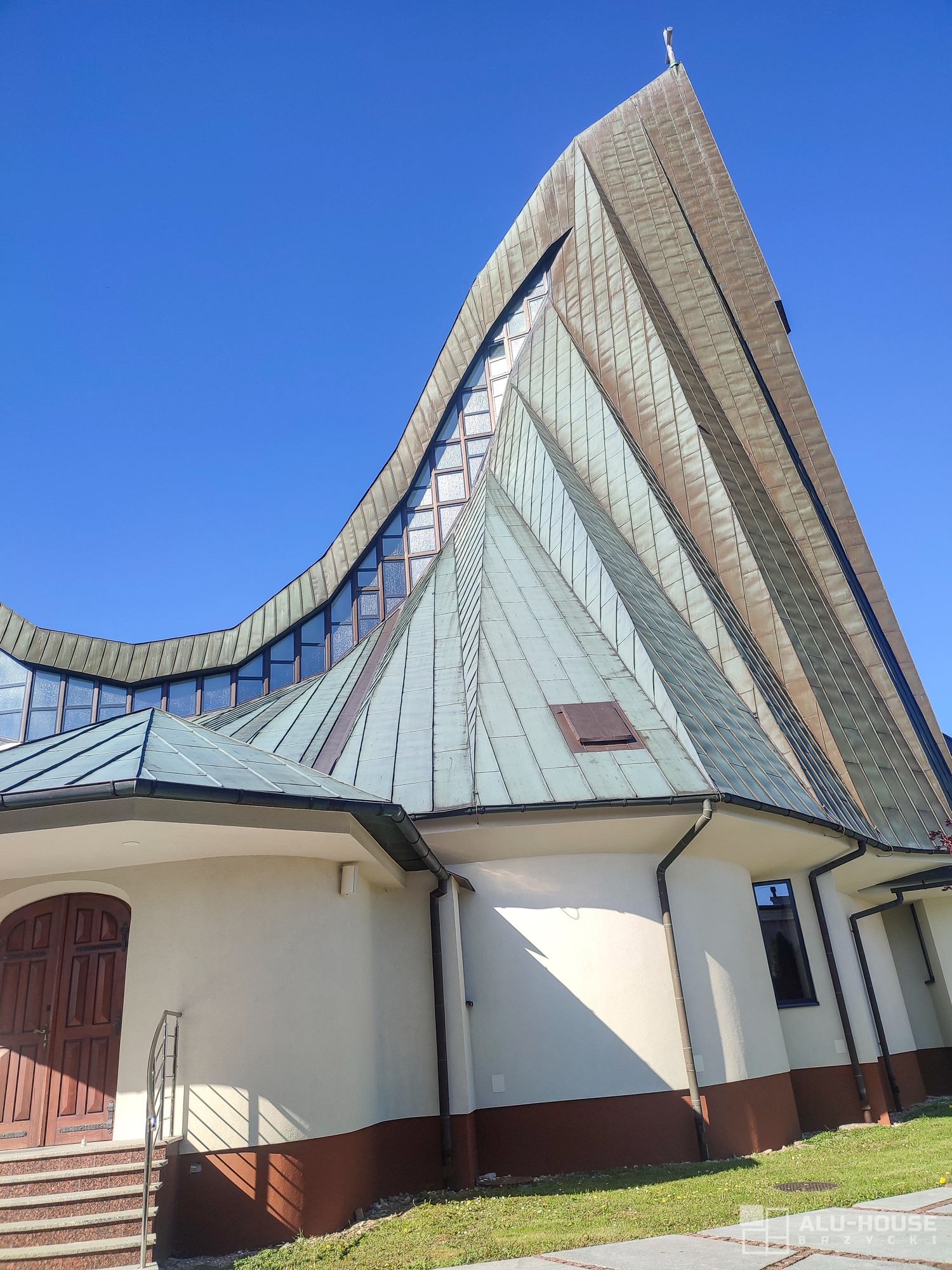 Nowoczesny budynek sakralny z innowacyjnym dachem z metalowych paneli, przeszklonymi lukarnami i drewnianymi drzwiami wejściowymi, widziany z dołu pod błękitnym niebem.