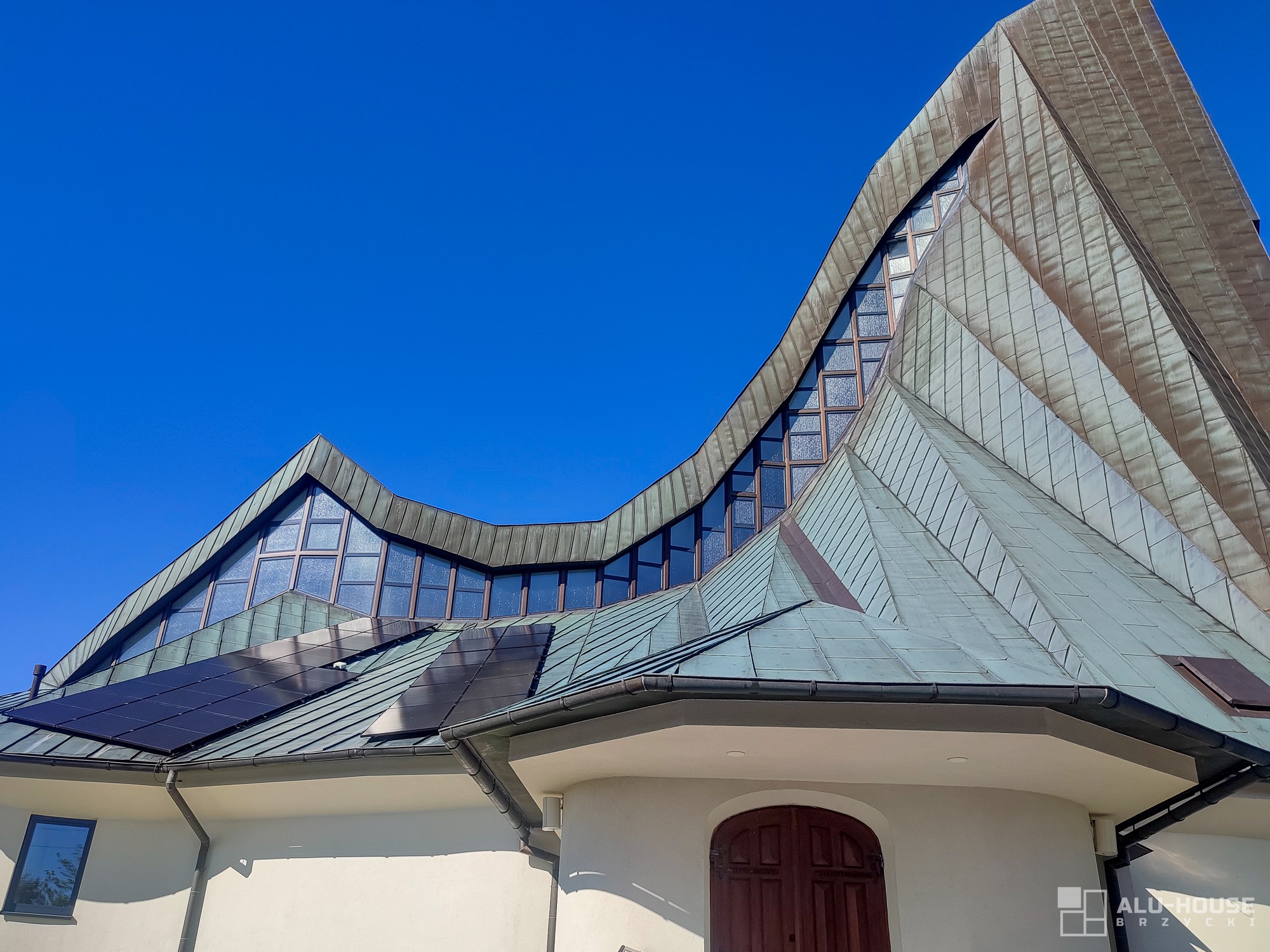 Nowoczesna bryła budynku z nietypowym dachem pokrytym metaliczną blachą i pasmem okien, panele słoneczne na dachu, błękitne niebo w tle.