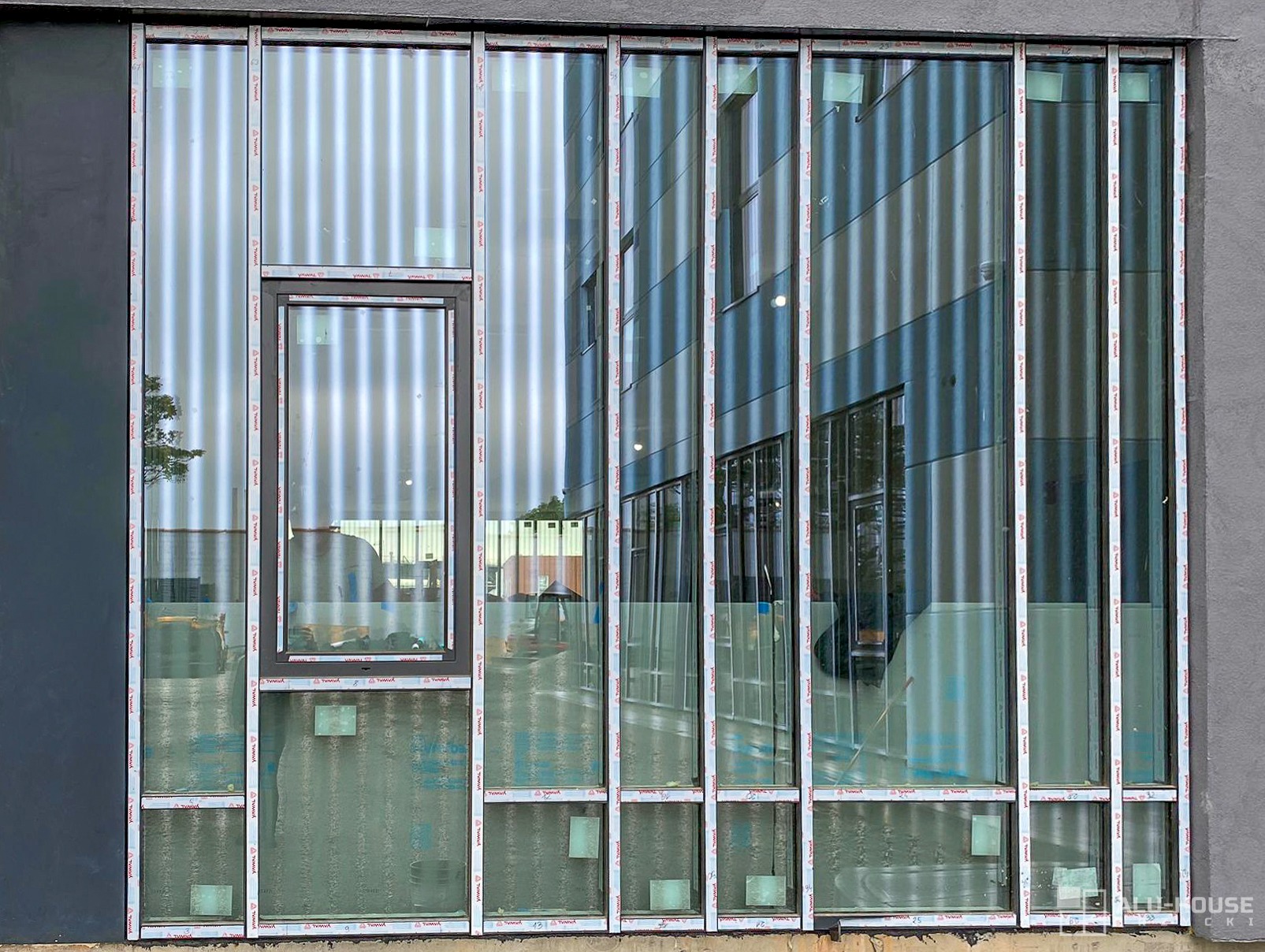 Fasada budynku z dużymi oknami z ramami z profili aluminiowych, odbijająca sąsiedni budynek z pionowymi panelami.