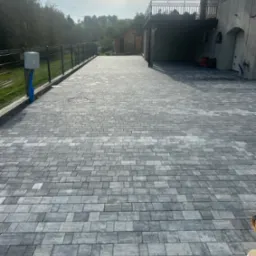 Ułożona szara kostka brukowa na podjeździe domu z balkonem, widoczna linia ogrodzenia i zieleń w tle.