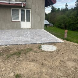 Mill-Bud Jakub Twaróg - Nowo ułożony, szary chodnik z kostki brukowej przylegający do ściany domu z balkonem, widoczny właz do studzienki i niezabezpieczone obrzeże z ziemi.