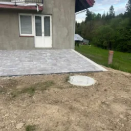 Nowo ułożony, szary chodnik z kostki brukowej przylegający do ściany domu z balkonem, widoczny właz do studzienki i niezabezpieczone obrzeże z ziemi.