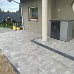 Mill-Bud Jakub Twaróg - Ułożona szara kostka brukowa przed budynkiem z kolumną, widoczna pompa ciepła, w tle domy i niebo.