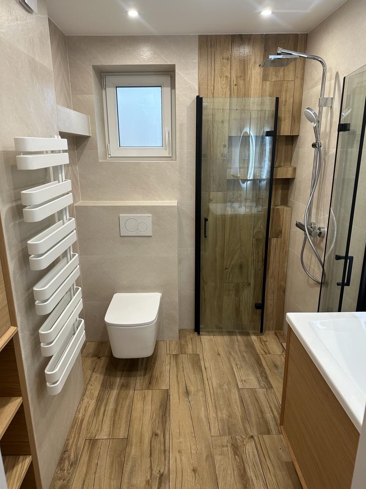 Nowoczesna łazienka z drewnopodobnymi płytkami, kabiną prysznicową typu walk-in z czarnymi profilami, wiszącą toaletą i grzejnikiem drabinkowym.