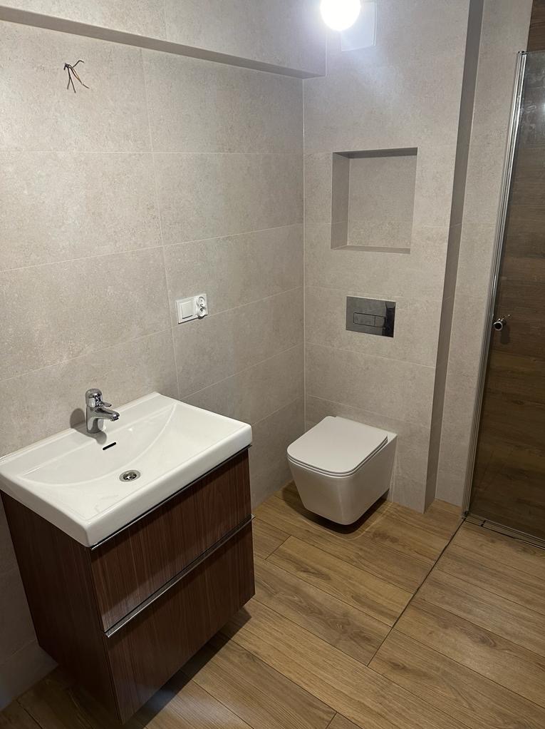Nowoczesna łazienka z beżowymi płytkami, drewnopodobną podłogą i wiszącą toaletą, umywalka z szafką w kolorze ciemnego drewna, minimalistyczna armatura.