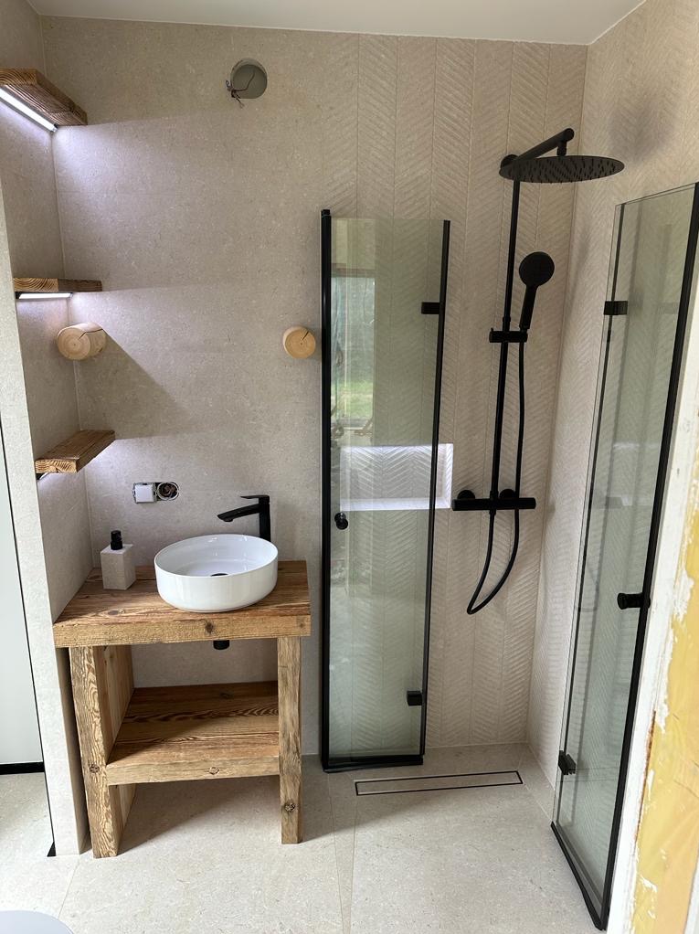 Nowoczesna łazienka z kabiną prysznicową typu walk-in z czarnymi profilami, umywalką nablatową na drewnianym blacie rustykalnym i drewnianymi półkami z podświetleniem LED, wykończona płytkami...