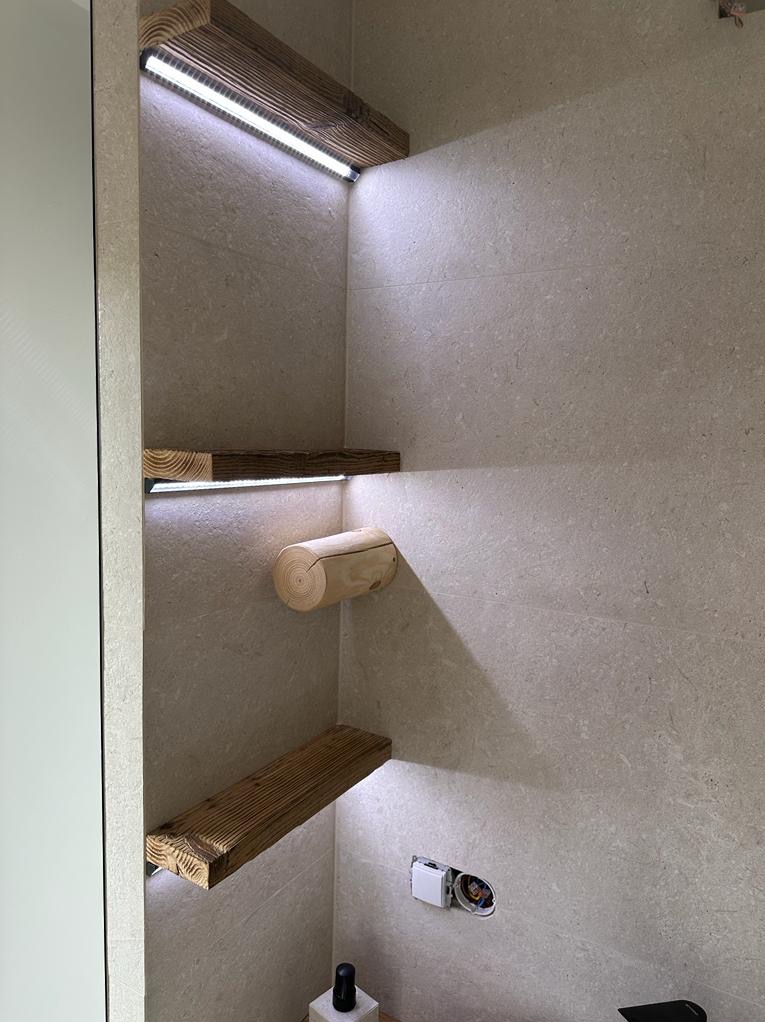 Narożna półka w łazience wykończona płytkami w odcieniach beżu, z trzema drewnianymi półkami podświetlonymi listwami LED, oraz uchwyt na papier toaletowy wykonany z kawałka pnia drzewa.