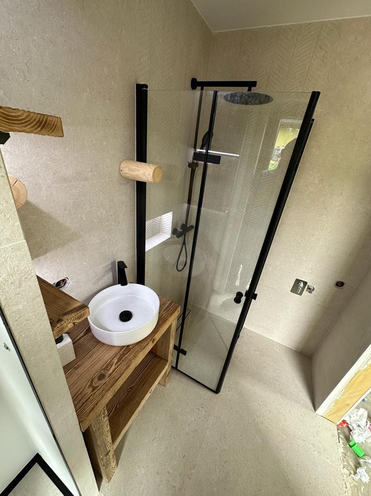 Nowoczesna łazienka z kabiną prysznicową z czarnymi profilami, umywalką nablatową na drewnianym blacie z półkami i drewnianymi akcentami na ścianie, wykończona jasnymi płytkami.