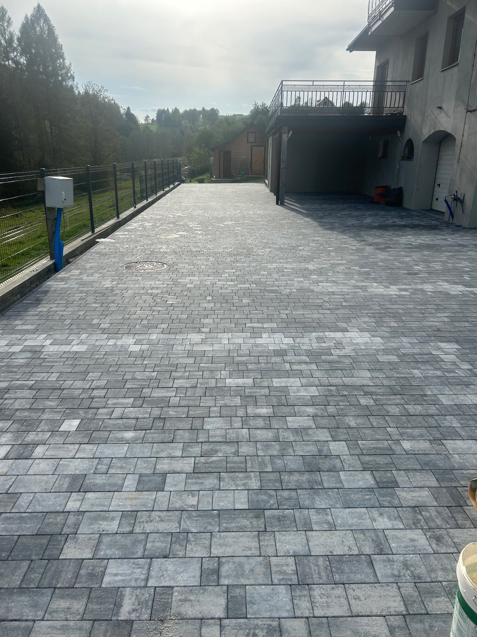 Ułożona szara kostka brukowa na podjeździe domu z balkonem, widoczna linia ogrodzenia i zieleń w tle.