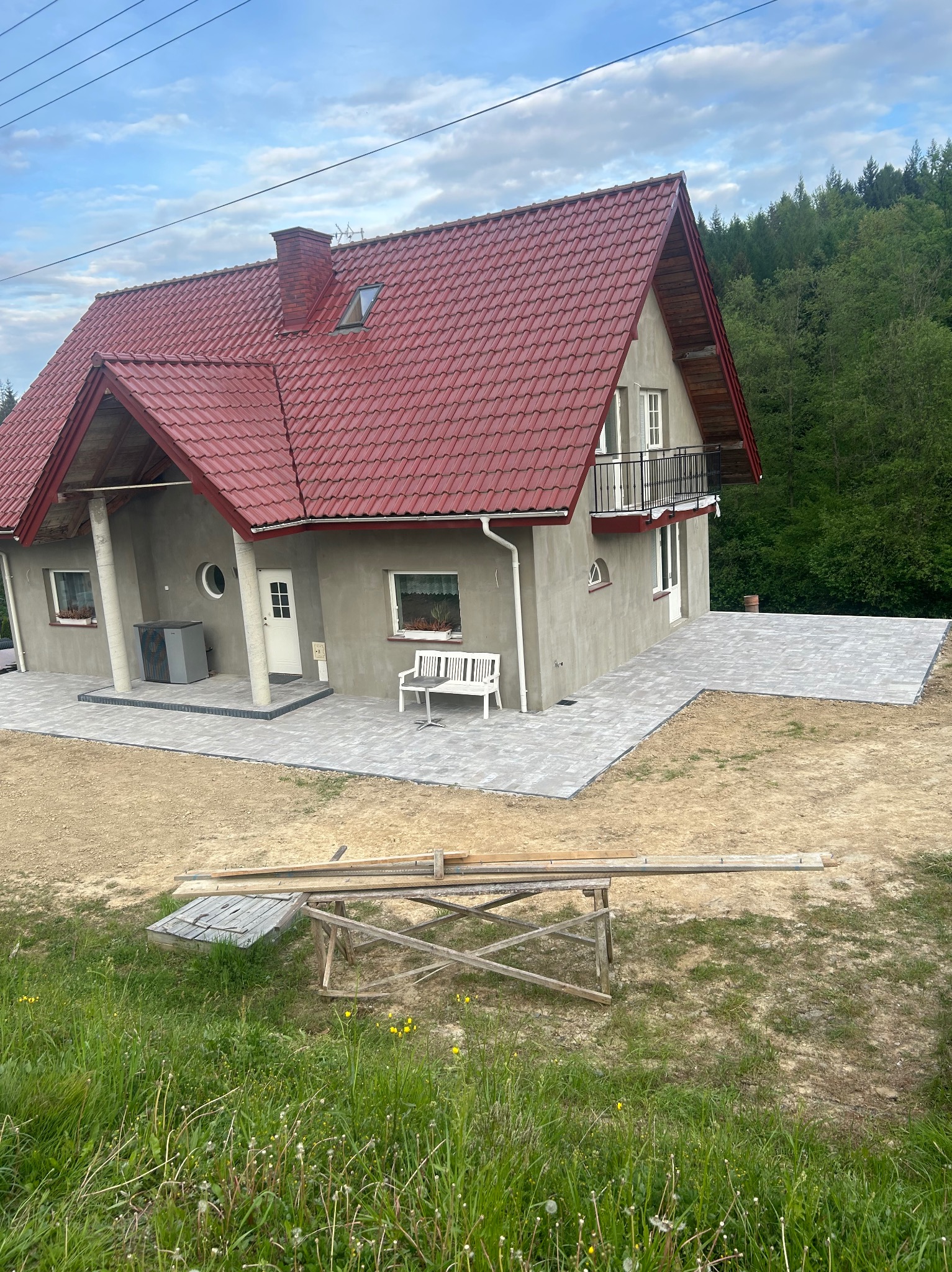 Nowo wykończony dom jednorodzinny z czerwoną dachówką i szarą elewacją, z ułożoną kostką brukową wokół, widok z perspektywy trawnika z drewnianym kozłem i deskami.