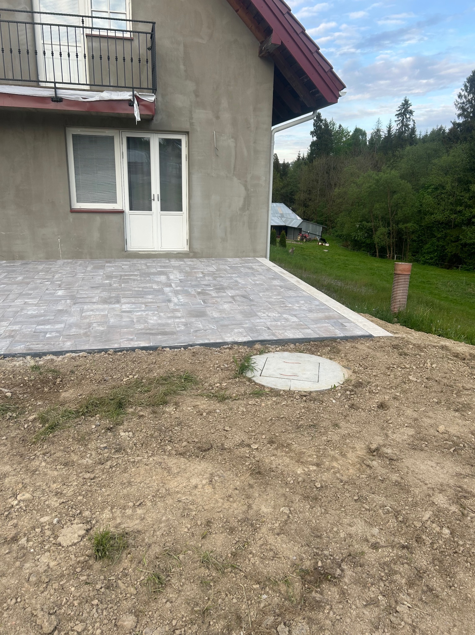 Nowo ułożony, szary chodnik z kostki brukowej przylegający do ściany domu z balkonem, widoczny właz do studzienki i niezabezpieczone obrzeże z ziemi.