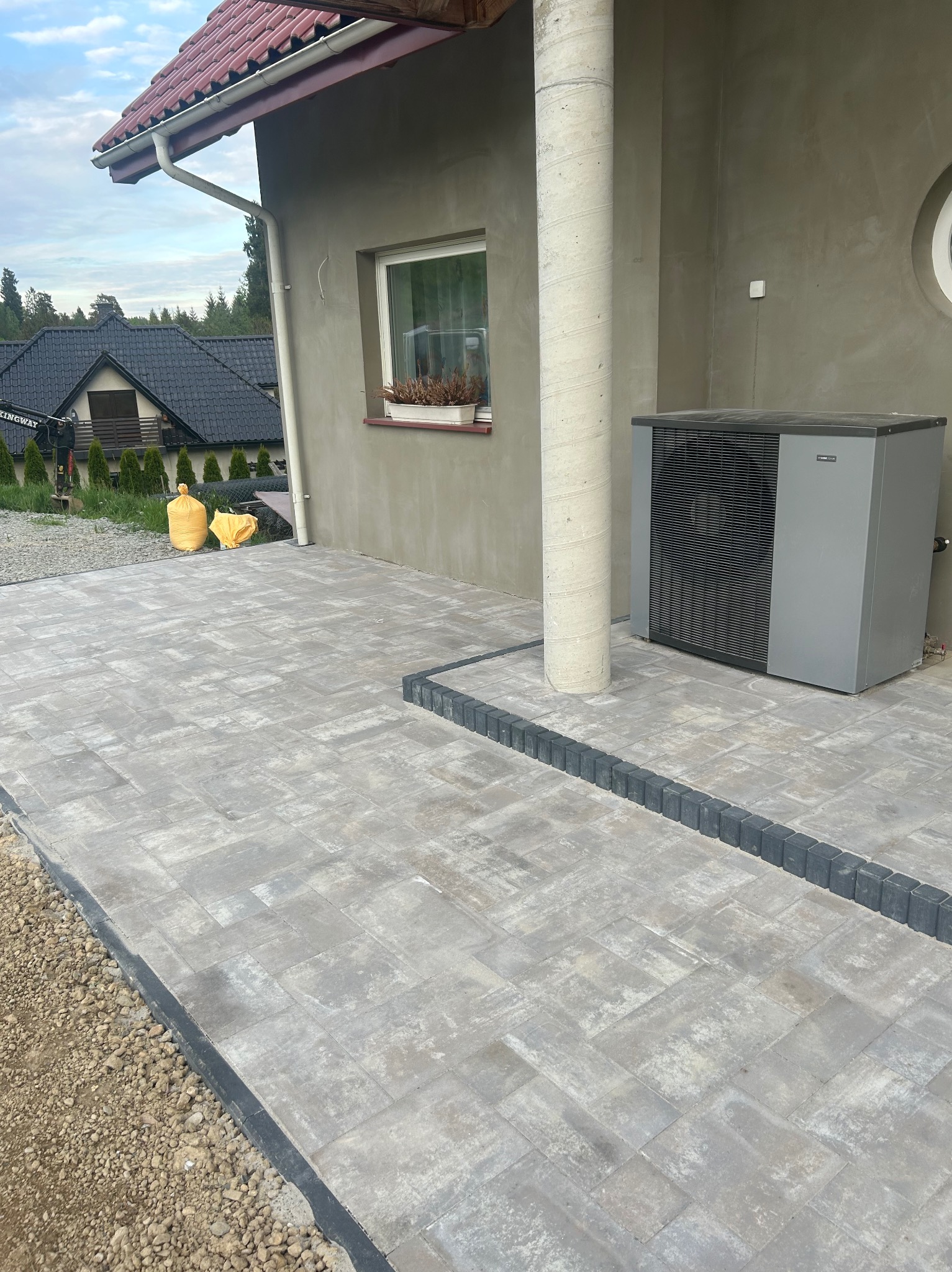 Ułożona szara kostka brukowa przed budynkiem z kolumną, widoczna pompa ciepła, w tle domy i niebo.