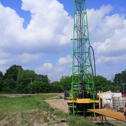 F.U.H. No-Kop Norbert Koprowski - Zielona wiertnica na podwoziu ciężarówki, widok z tyłu, rury wiertnicze na stojaku, błękitne niebo z obłokami w tle.