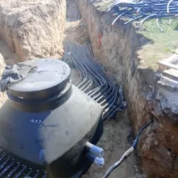 Czarny zbiornik w wykopie ziemnym, z licznymi rurami wchodzącymi do niego, widoczne połączenia i fragmenty instalacji, ziemia piaszczysta.