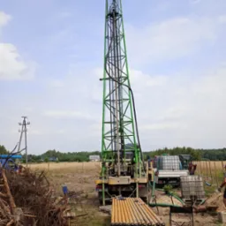 Wiertnica na podwoziu samochodowym z zieloną kratownicą masztu, rury wiertnicze ułożone na kozłach, zbiorniki na wodę w tle, teren budowy z wykopaną ziemią.