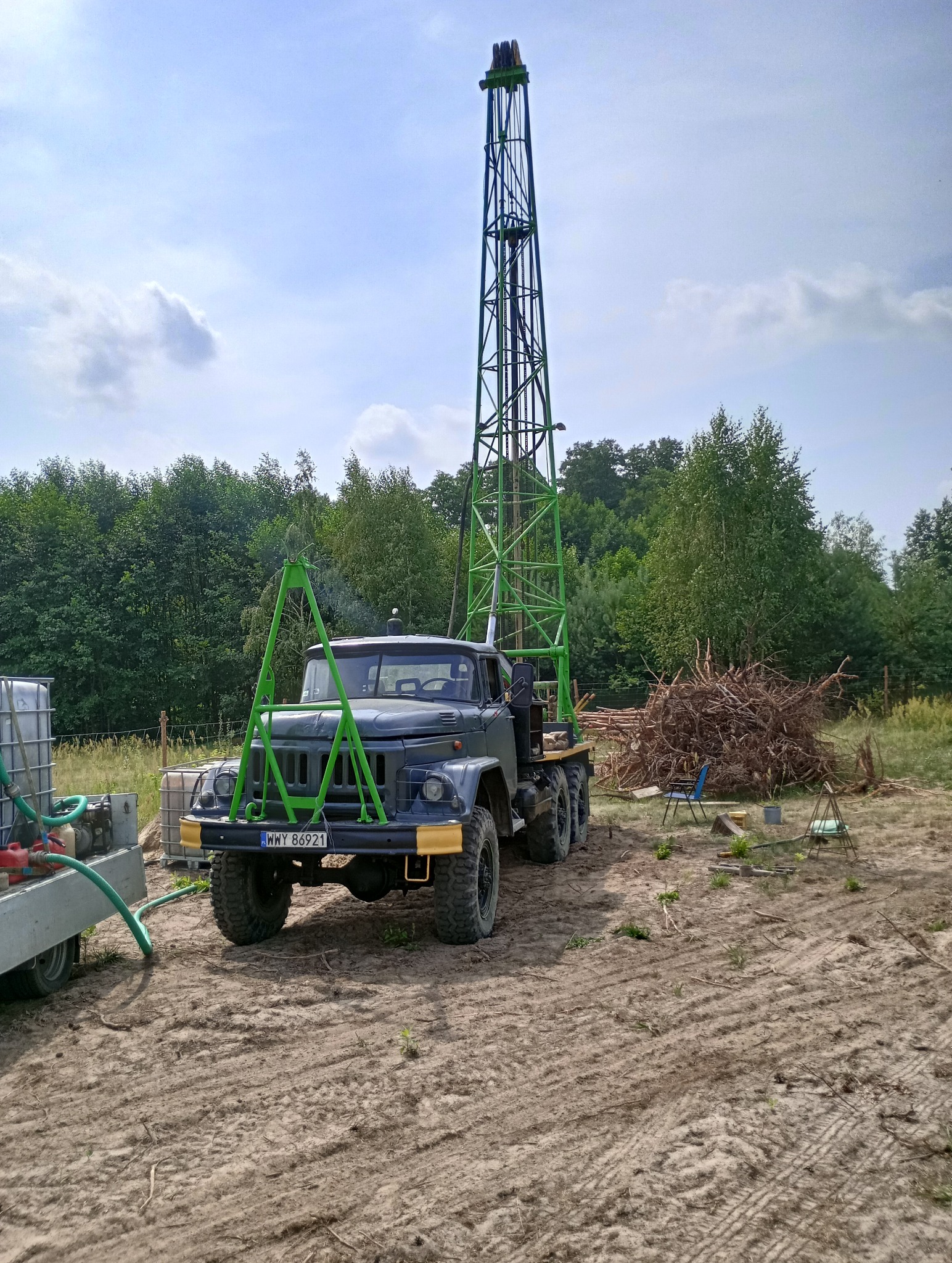 Ciężarówka wiertnicza z wysoką wieżą wiertniczą, zaparkowana na piaszczystym terenie z widocznym stosem gałęzi i zbiornikami wody w tle, pod błękitnym niebem z chmurami.