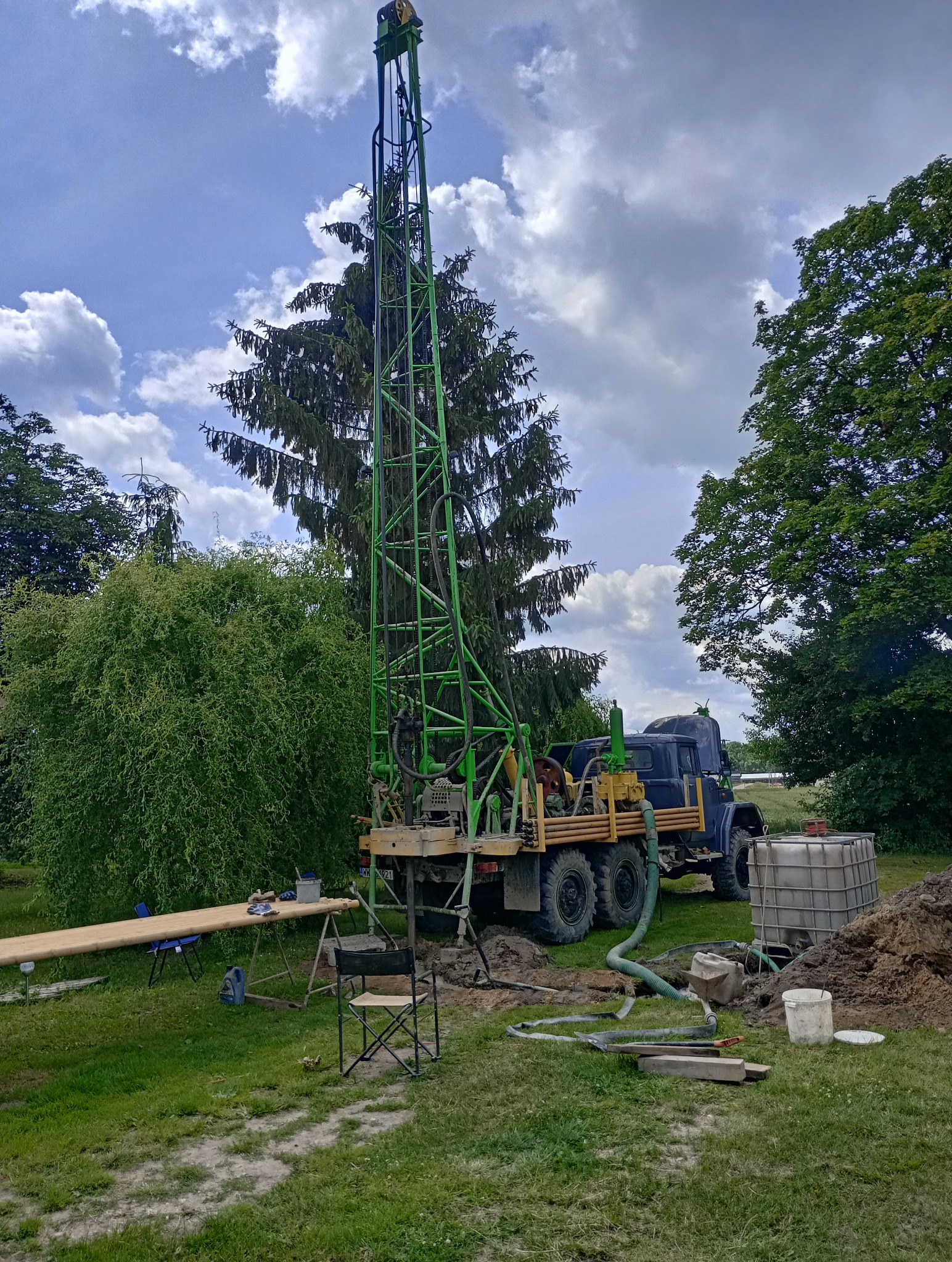 Wiertnica zamontowana na ciężarówce, z zieloną kratownicową wieżą, podczas pracy na zewnątrz w otoczeniu drzew; widoczny wykop, węże i zbiornik na wodę.