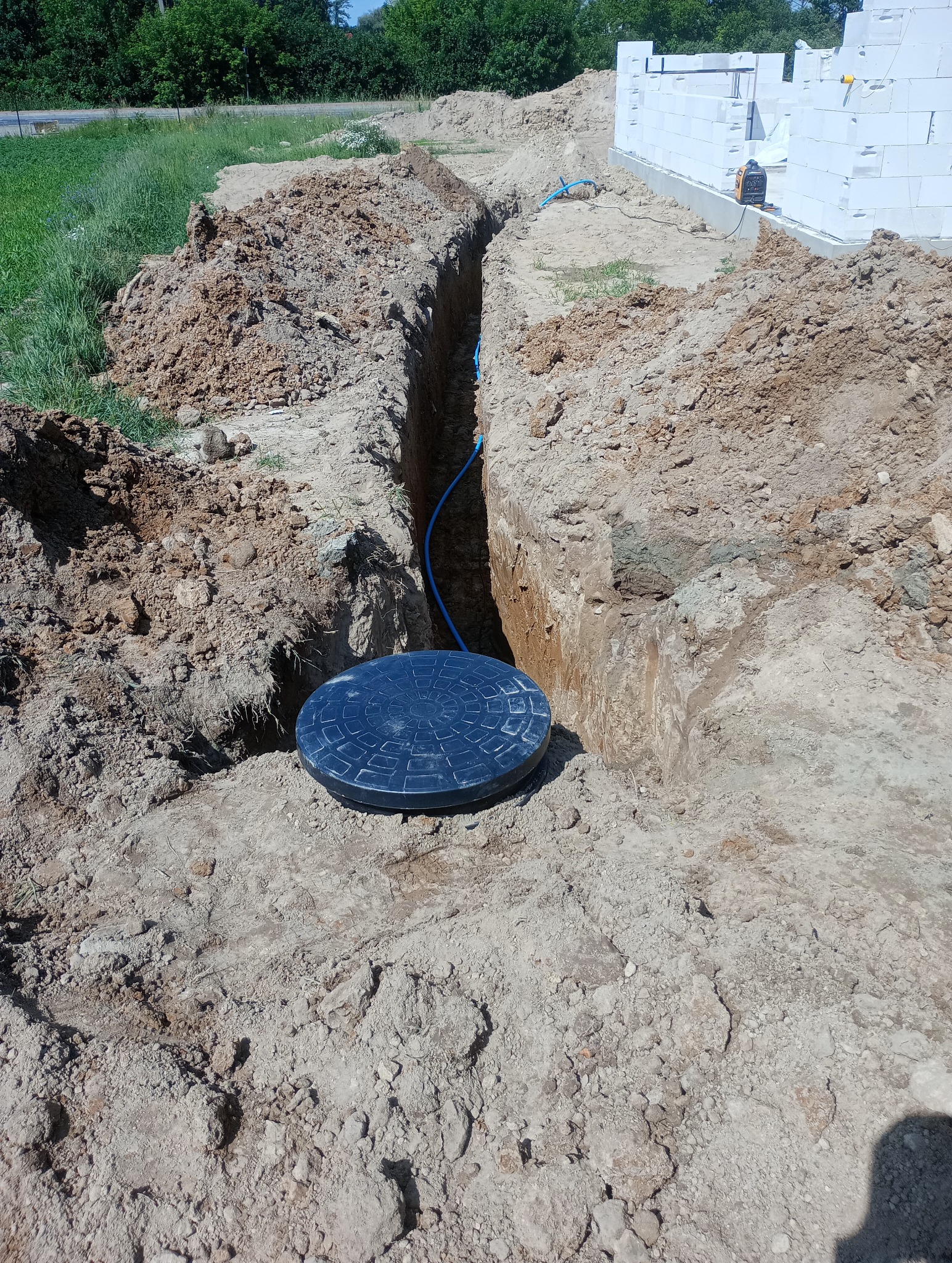 Wykopany wąski rów w ziemi z niebieską rurą i czarną pokrywą studzienki, obok fundamentów budynku z białego pustaka.