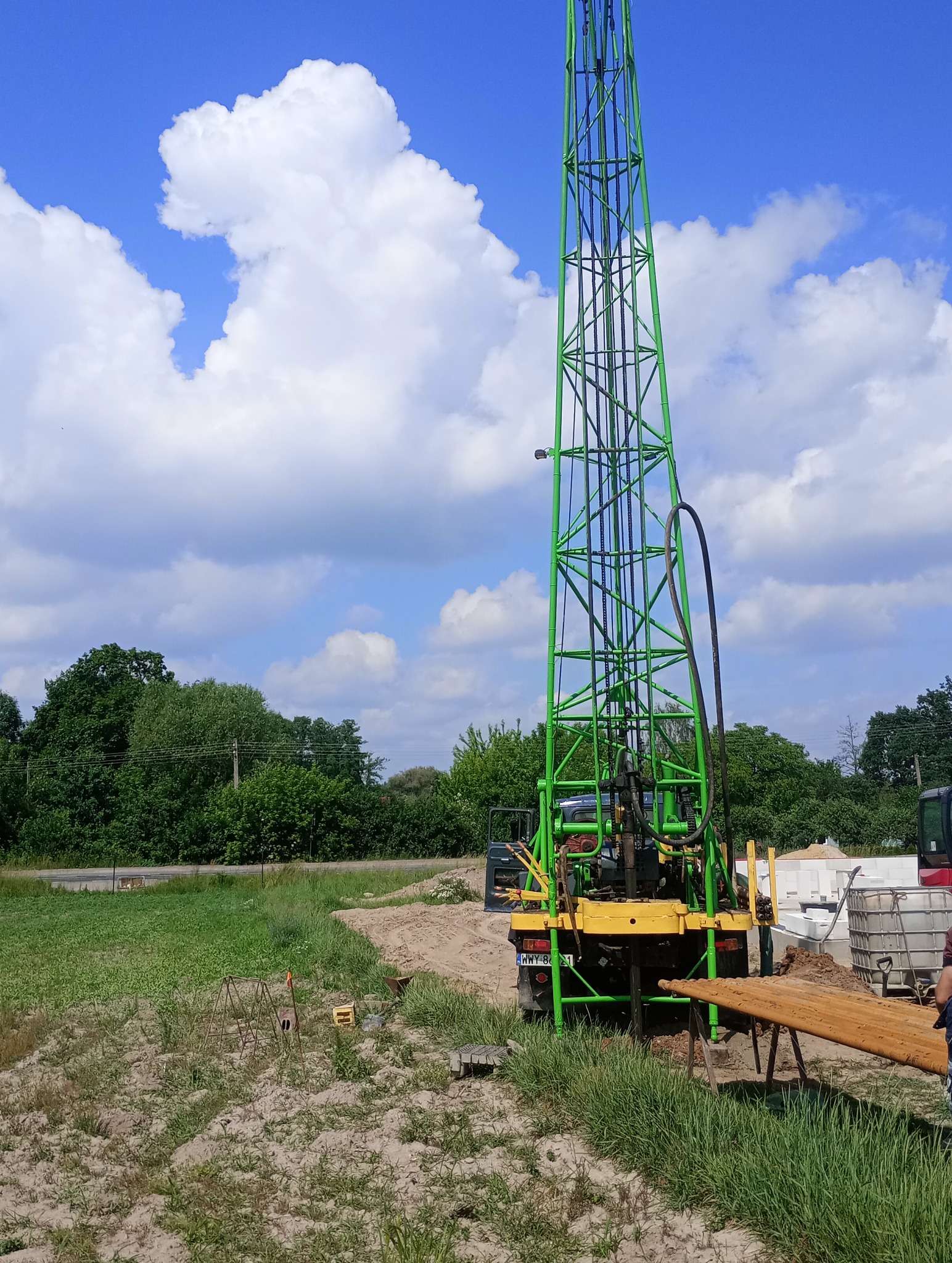 Zielona wiertnica na podwoziu ciężarówki, widok z tyłu, rury wiertnicze na stojaku, błękitne niebo z obłokami w tle.