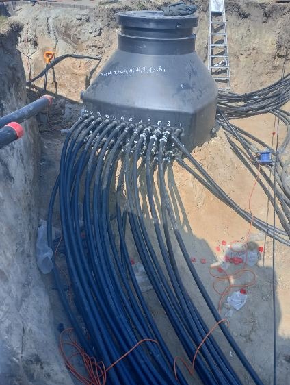 Czarna, plastikowa studzienka rozdzielcza kabli energetycznych w wykopie ziemnym, z licznymi, grubymi kablami wchodzącymi do niej. Widoczne oznaczenia numerów na studzience i drabina oparta...