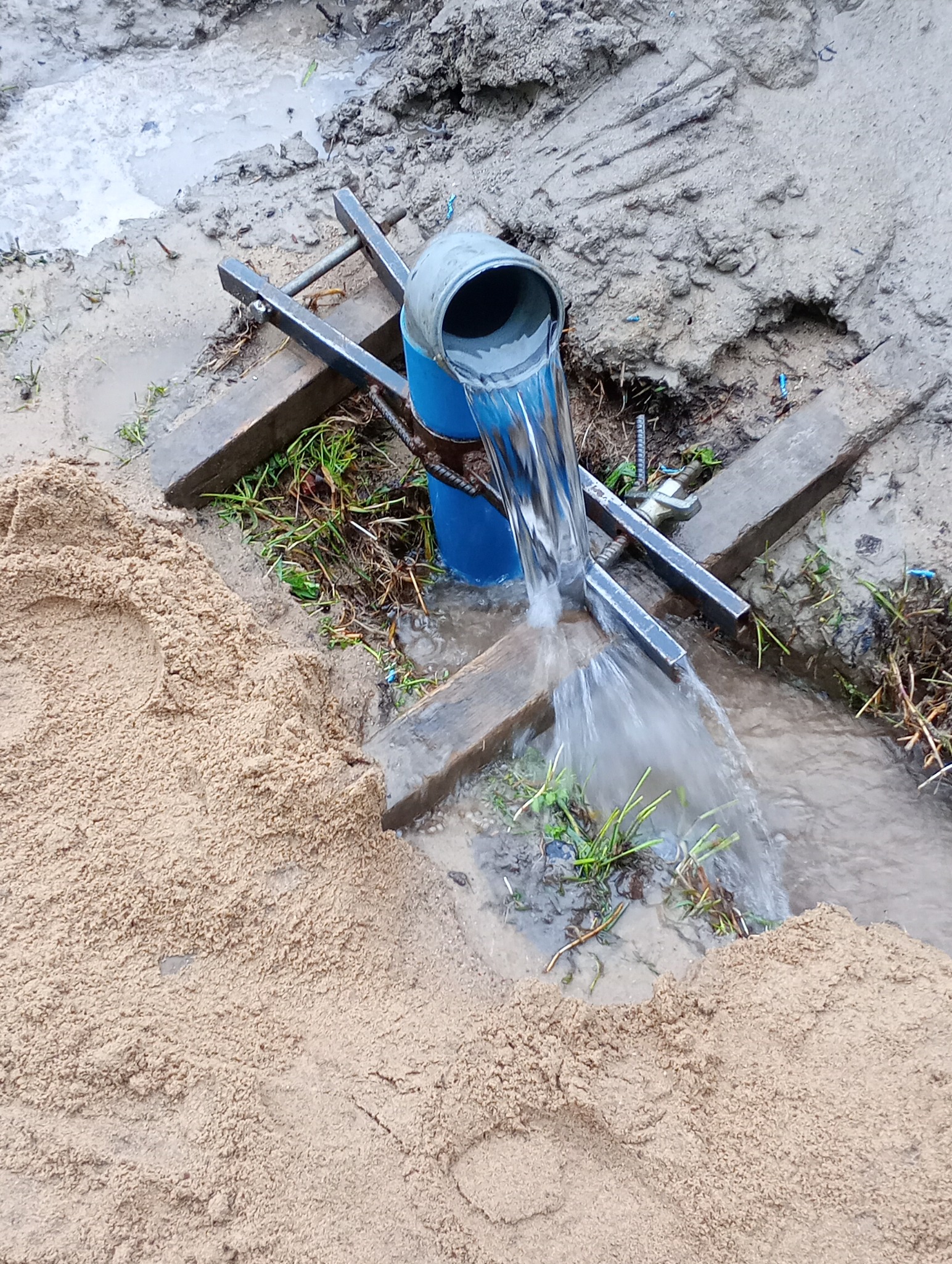 Woda wypływająca z niebieskiej rury w ziemi, otoczona piaskiem i błotem, wsparta na metalowej konstrukcji.