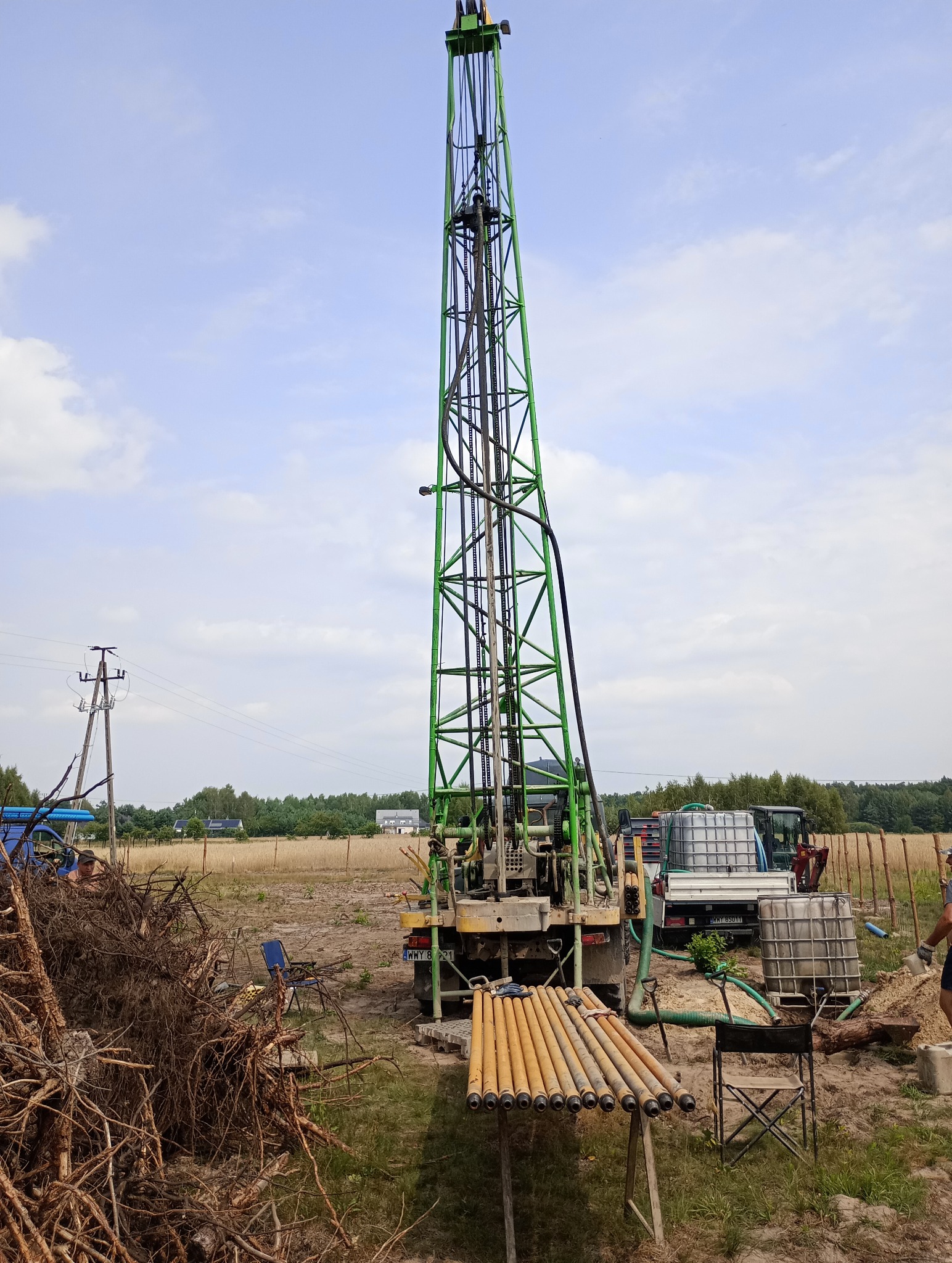 Wiertnica na podwoziu samochodowym z zieloną kratownicą masztu, rury wiertnicze ułożone na kozłach, zbiorniki na wodę w tle, teren budowy z wykopaną ziemią.