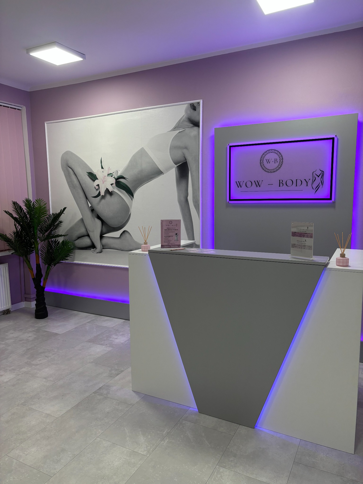 Recepcja w gabinecie z podświetlanym logo WOW-BODY, plakatem kobiety z kwiatami i dekoracyjną palmą w tle, całość utrzymana w odcieniach szarości i fioletu.