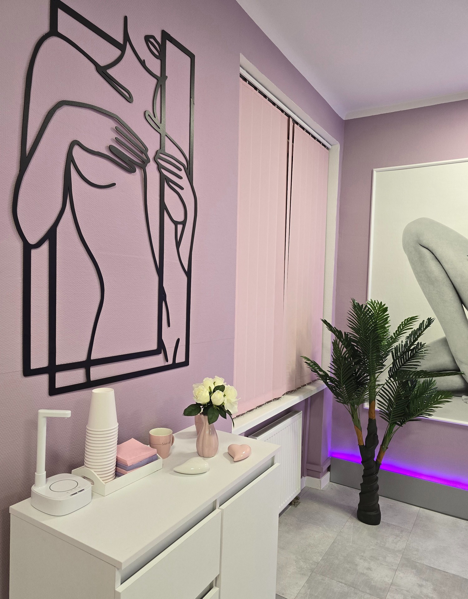 Nowoczesny gabinet kosmetyczny w odcieniach fioletu z minimalistyczną dekoracją ścienną przedstawiającą kobiecą sylwetkę, sztuczną palmą i białymi meblami.