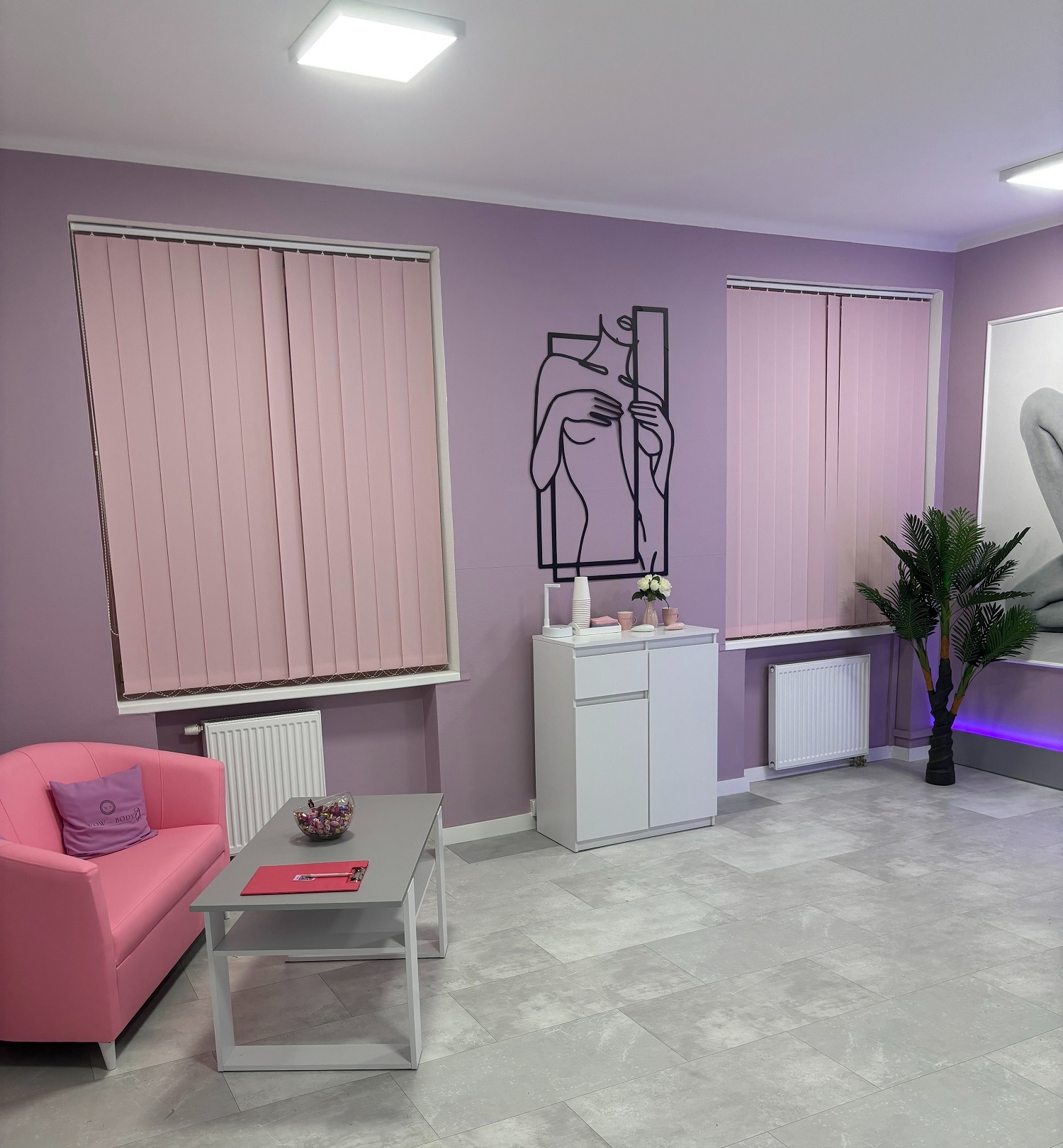Jasne wnętrze gabinetu kosmetycznego z fioletowymi ścianami, różowymi zasłonami i sofą, minimalistyczną dekoracją ścienną, szafką z kosmetykami i sztuczną palmą.