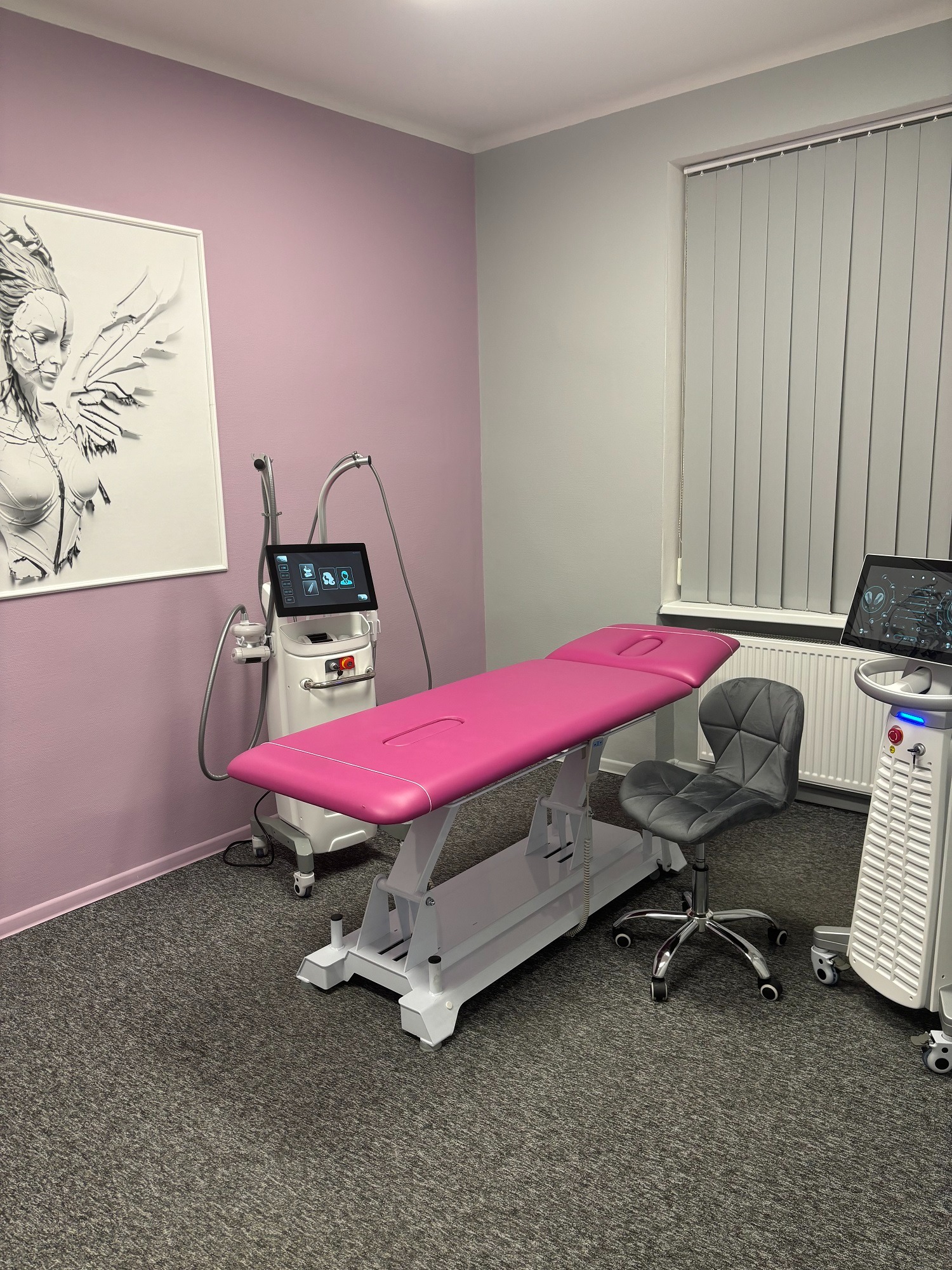 Pokój zabiegowy z różowym leżanką, dwoma urządzeniami kosmetycznymi z ekranami dotykowymi, szarym krzesłem obrotowym i grafiką kobiety ze skrzydłami na ścianie.