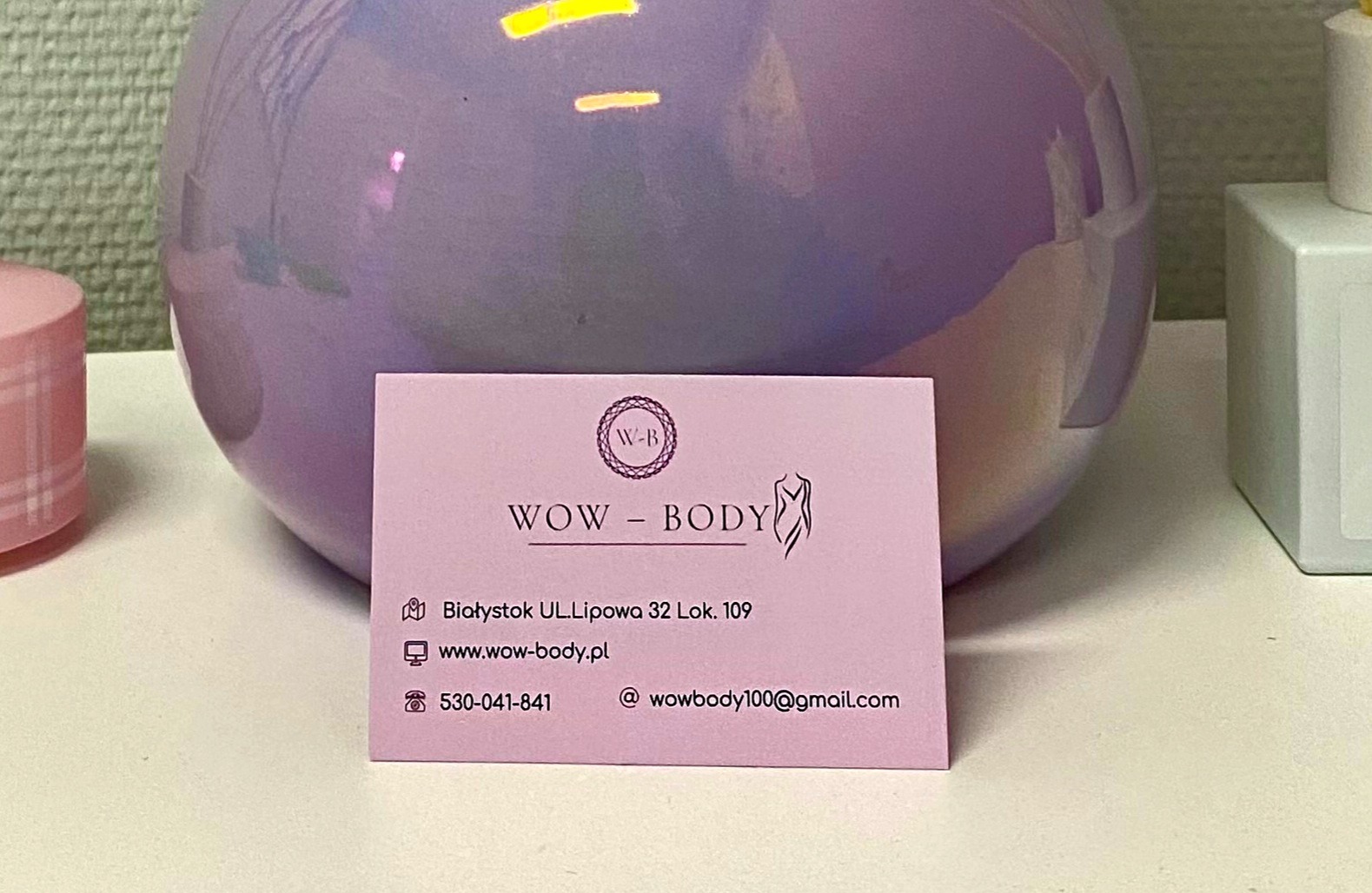 Wizytówka firmy WOW BODY z adresem w Białymstoku na tle liliowej, okrągłej lampy i białego flakonu.