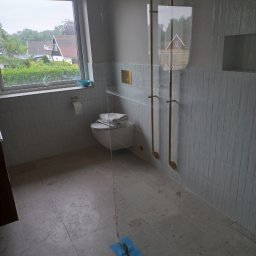 Dawbud - Nowoczesna łazienka z szarą, pionowo ułożoną glazurą, szklaną kabiną prysznicową z złotymi relingami, toaletą z ręcznikami, oknem z widokiem na domy i ogród, oraz kamienną podłogą z niebieską taśmą...