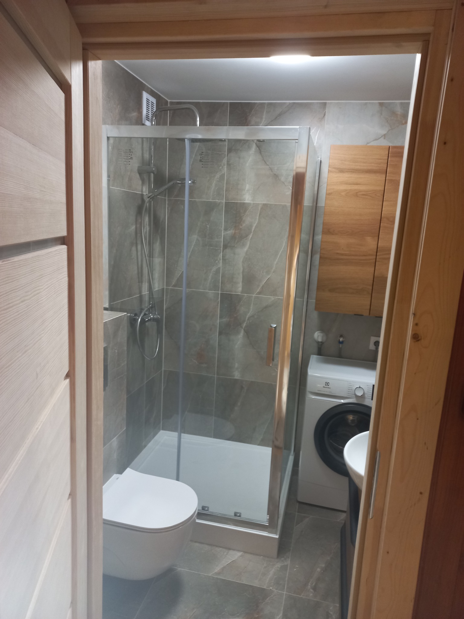 Nowoczesna łazienka z kabiną prysznicową, umywalką, toaletą i pralką, wykończona płytkami imitującymi marmur i drewnianymi elementami, widziana zza drewnianej ościeżnicy.