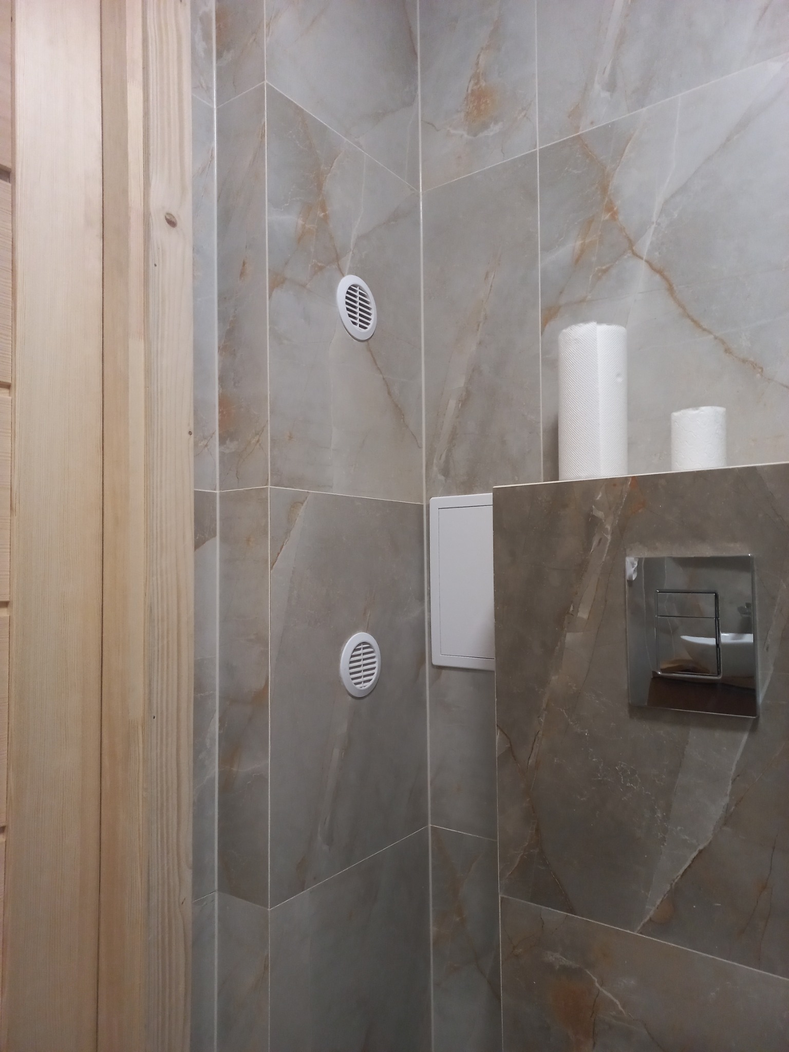 Wykończona łazienka z marmurowymi płytkami, ukrytym systemem spłukiwania WC, dwoma rolkami papieru toaletowego i drewnianym wykończeniem drzwi.