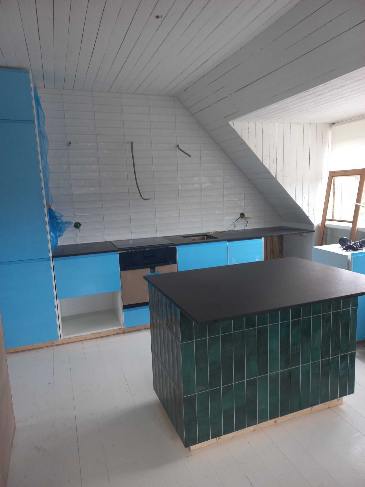 Kuchnia w trakcie prac wykończeniowych: niebieskie szafki z czarnym blatem, wyspa z ciemnozielonych płytek, białe płytki na ścianie pod skosem i drewniana podłoga pomalowana na biało.