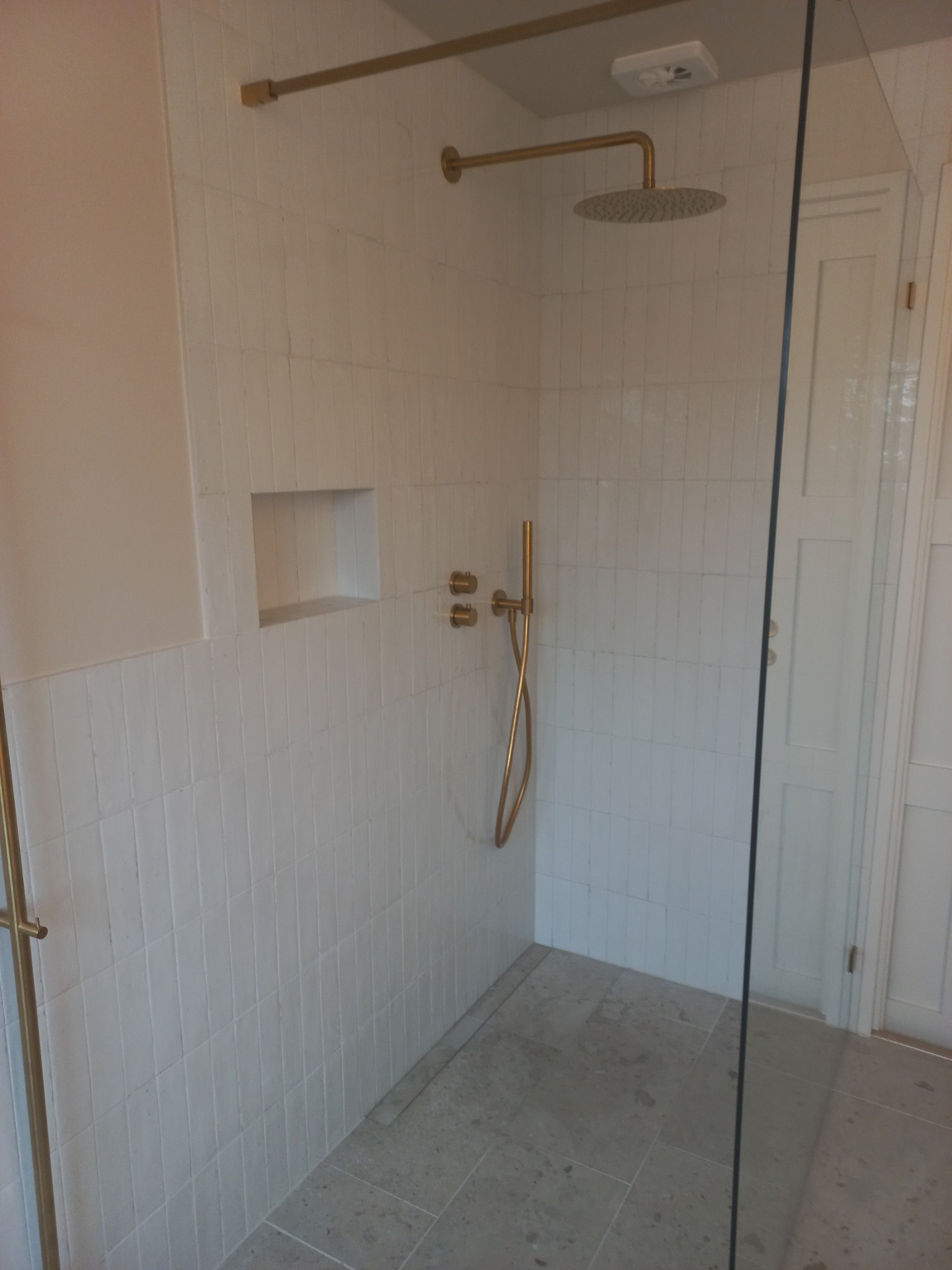 Wykończona kabina prysznicowa walk-in z białymi, wąskimi płytkami ułożonymi pionowo i złotą armaturą w Jaworznie. Widoczna wnęka w ścianie wyłożona płytkami, szklana ściana kabiny oraz kamienna...