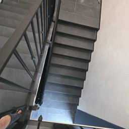 Damian Tyl - Schody wyłożone szarymi płytkami, z czarną metalową balustradą, widok z góry, częściowo widoczne stopy w klapkach, w tle wiadro i worki budowlane.