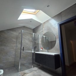 Damian Tyl - Nowoczesna łazienka na poddaszu z kabiną prysznicową, okrągłym lustrem z podświetleniem LED, czarną szafką pod umywalką i oknem dachowym.