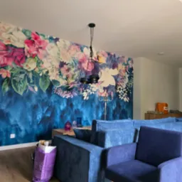 Salon z fototapetą w kwiaty na niebieskim tle, oświetlony nowoczesną lampą wiszącą i świecznikiem, z widoczną częścią niebieskiej sofy i fotelem.