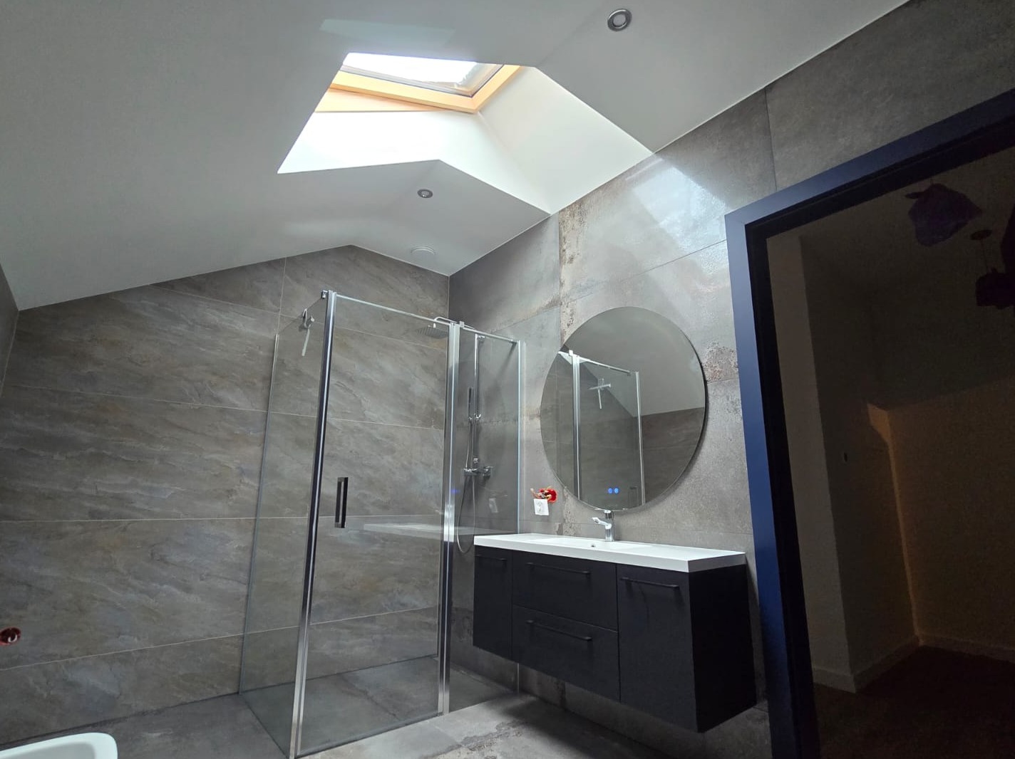 Nowoczesna łazienka na poddaszu z oknem dachowym, kabiną prysznicową ze szklanymi drzwiami, okrągłym lustrem z podświetleniem LED nad czarną szafką z umywalką.