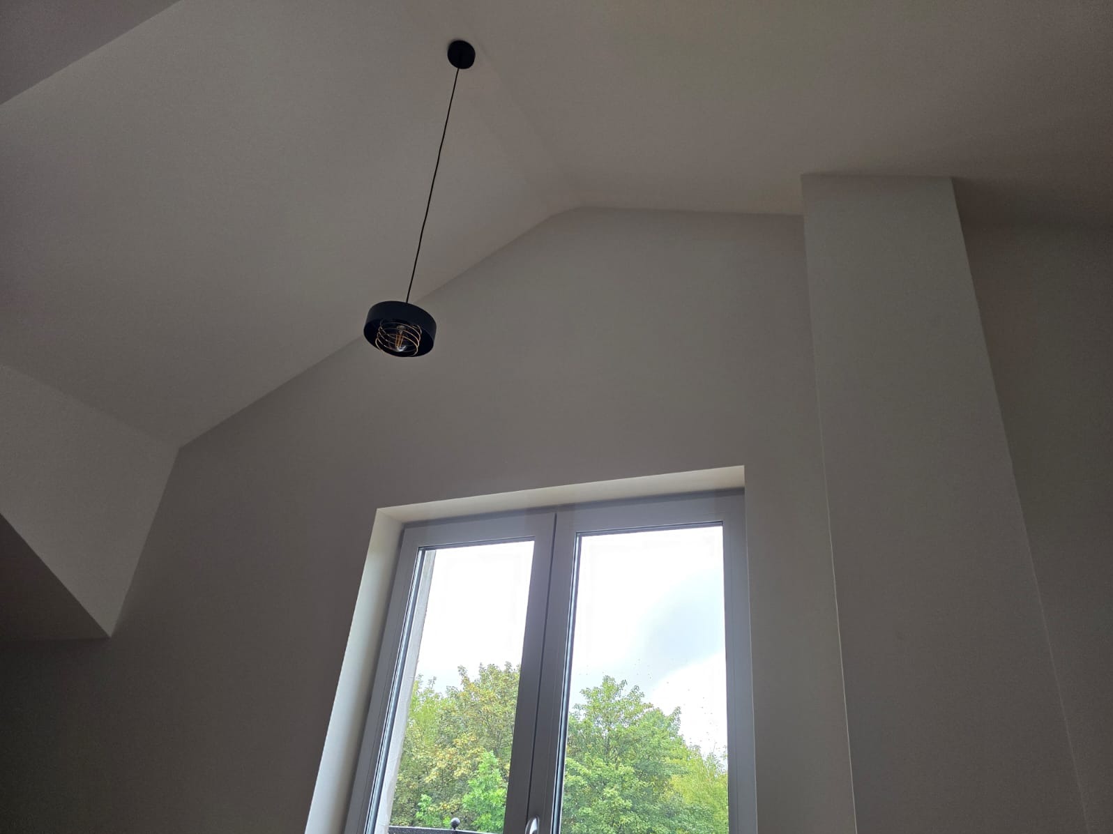 Nowoczesna czarna lampa wisząca z dekoracyjnym żarnikiem na tle białego, świeżo pomalowanego sufitu i ściany z oknem z widokiem na drzewa.