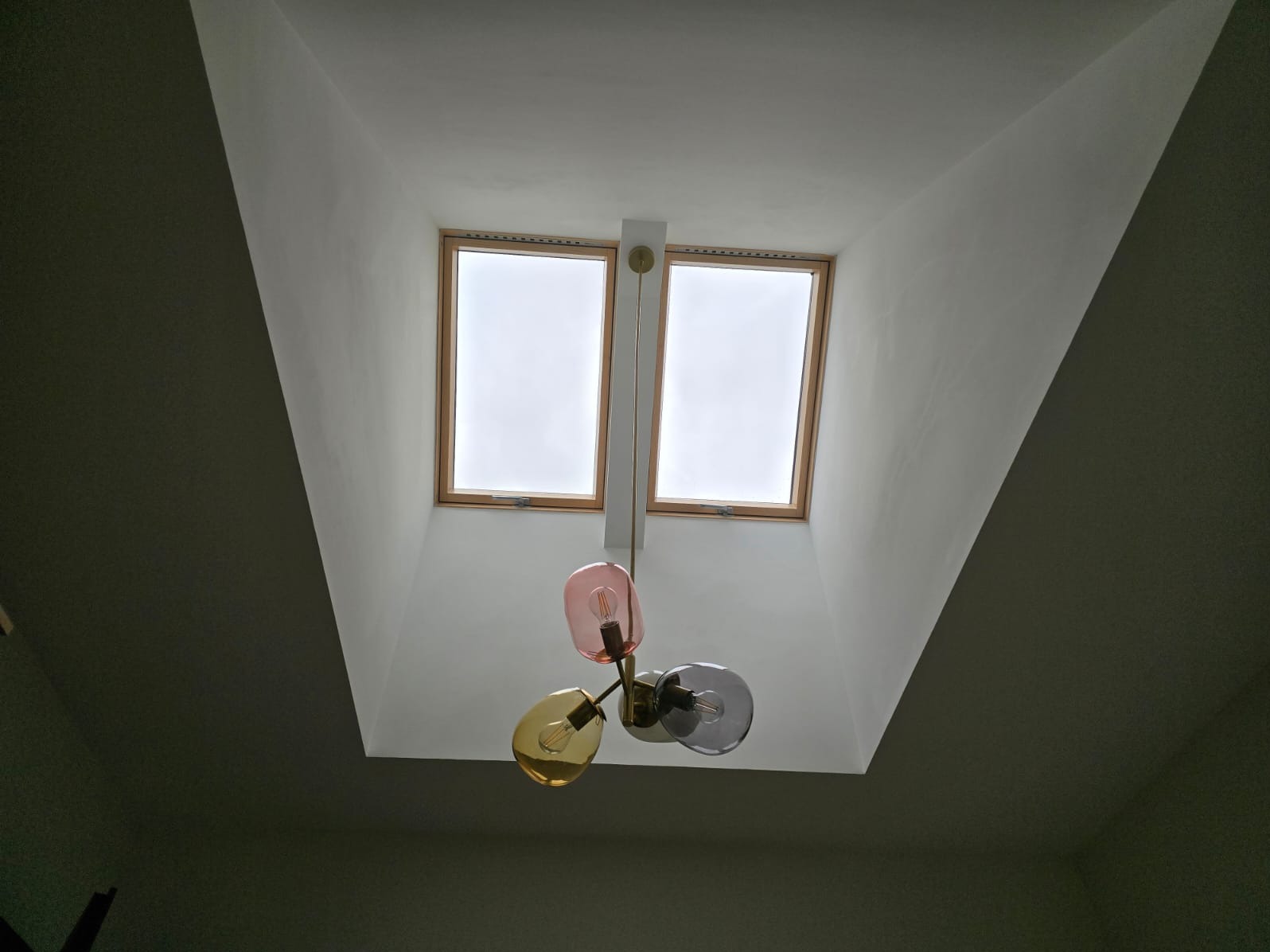 Dwa okna dachowe w białym suficie pod skosem, poniżej designerska lampa z trzema szklanymi kloszami w kolorach: różowym, żółtym i szarym.