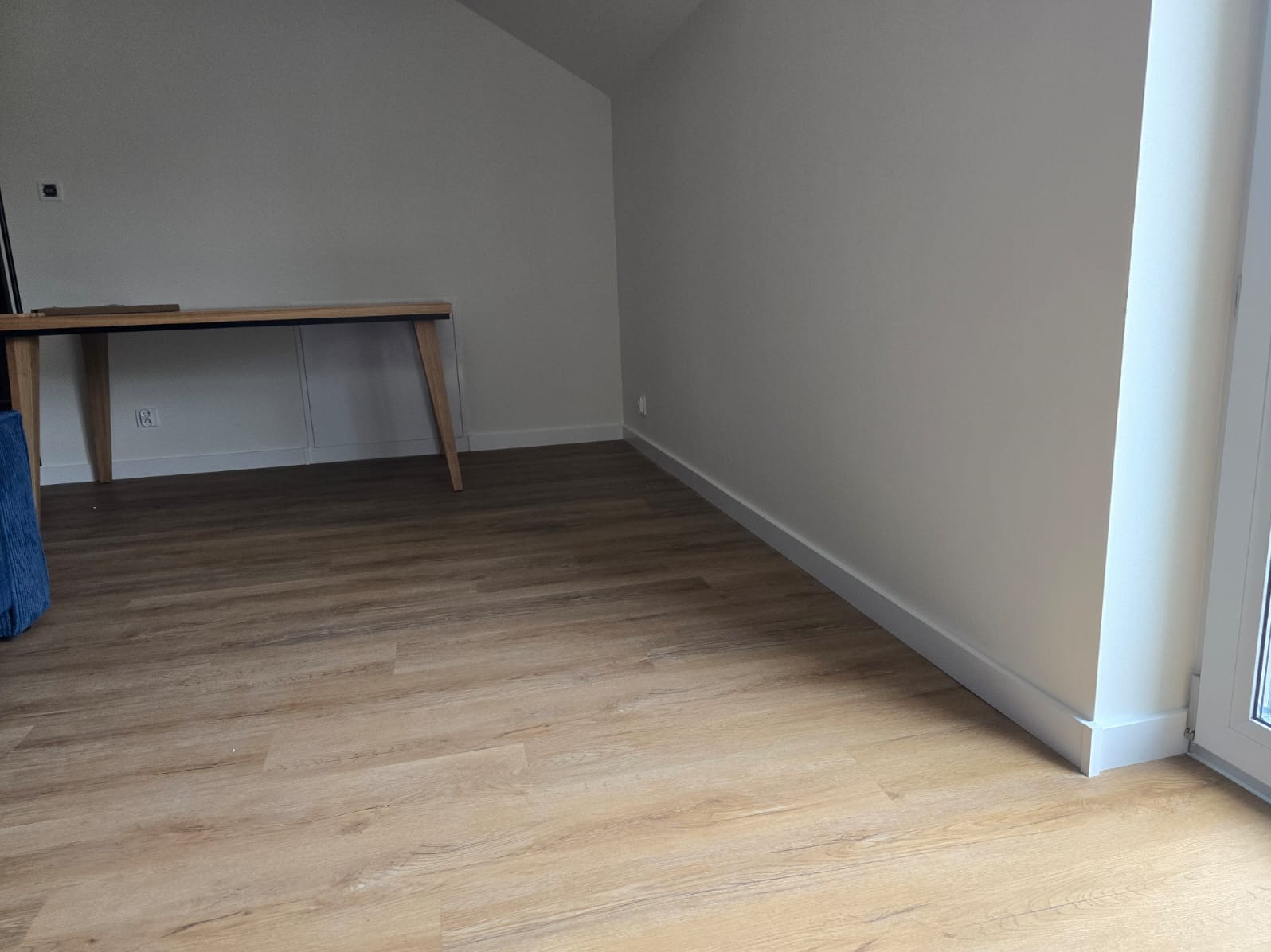 Jasne panele podłogowe imitujące drewno w nowo wykończonym pokoju z białymi ścianami i listwami przypodłogowymi, widoczny fragment biurka.