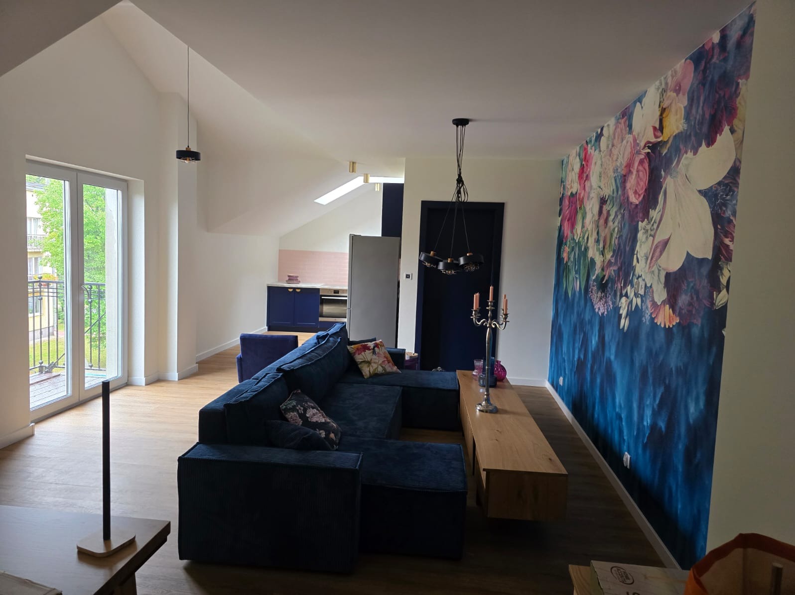 Jasne wnętrze mieszkania z niebieską sofą narożną, drewnianą podłogą, ścianą z motywem kwiatowym i aneksem kuchennym w tle