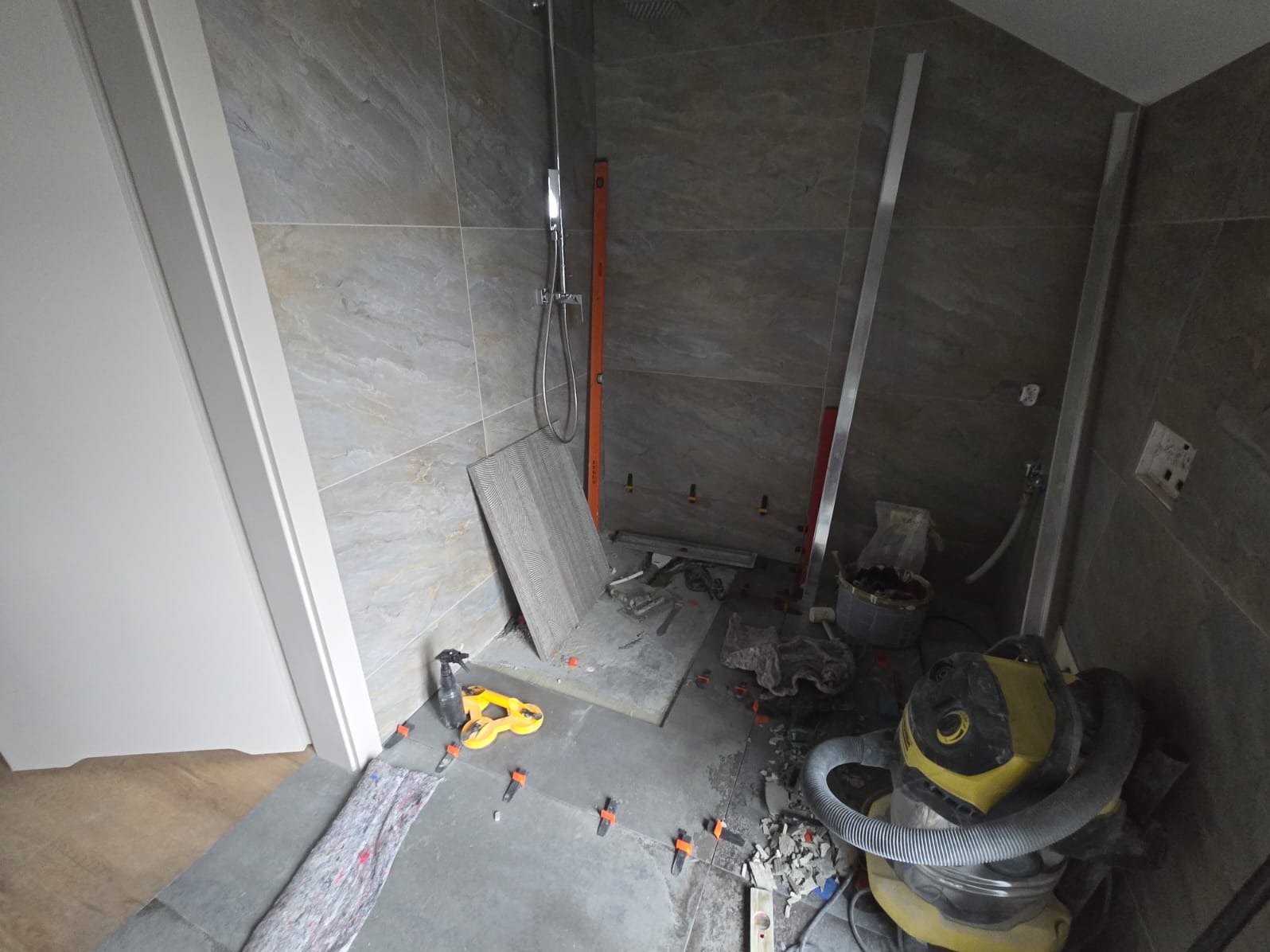 Montaż płytek w łazience: widok na podłogę z szarymi płytkami, poziomica, odkurzacz przemysłowy i narzędzia glazurnicze podczas układania płytek.